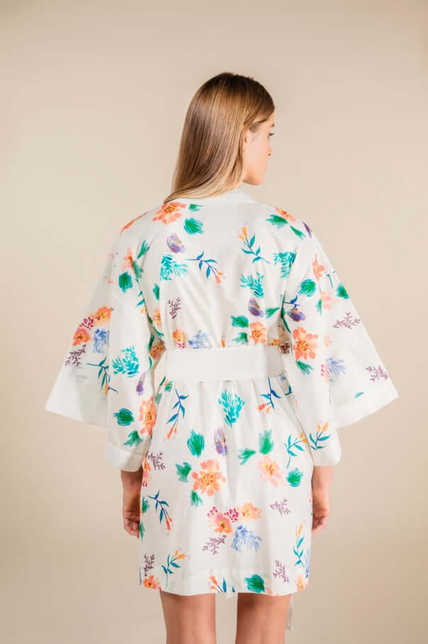 Back view of a woman wearing a floral Jasmine Cotton Robe made of 100% cotton, featuring vibrant flowers on ivory fabric.